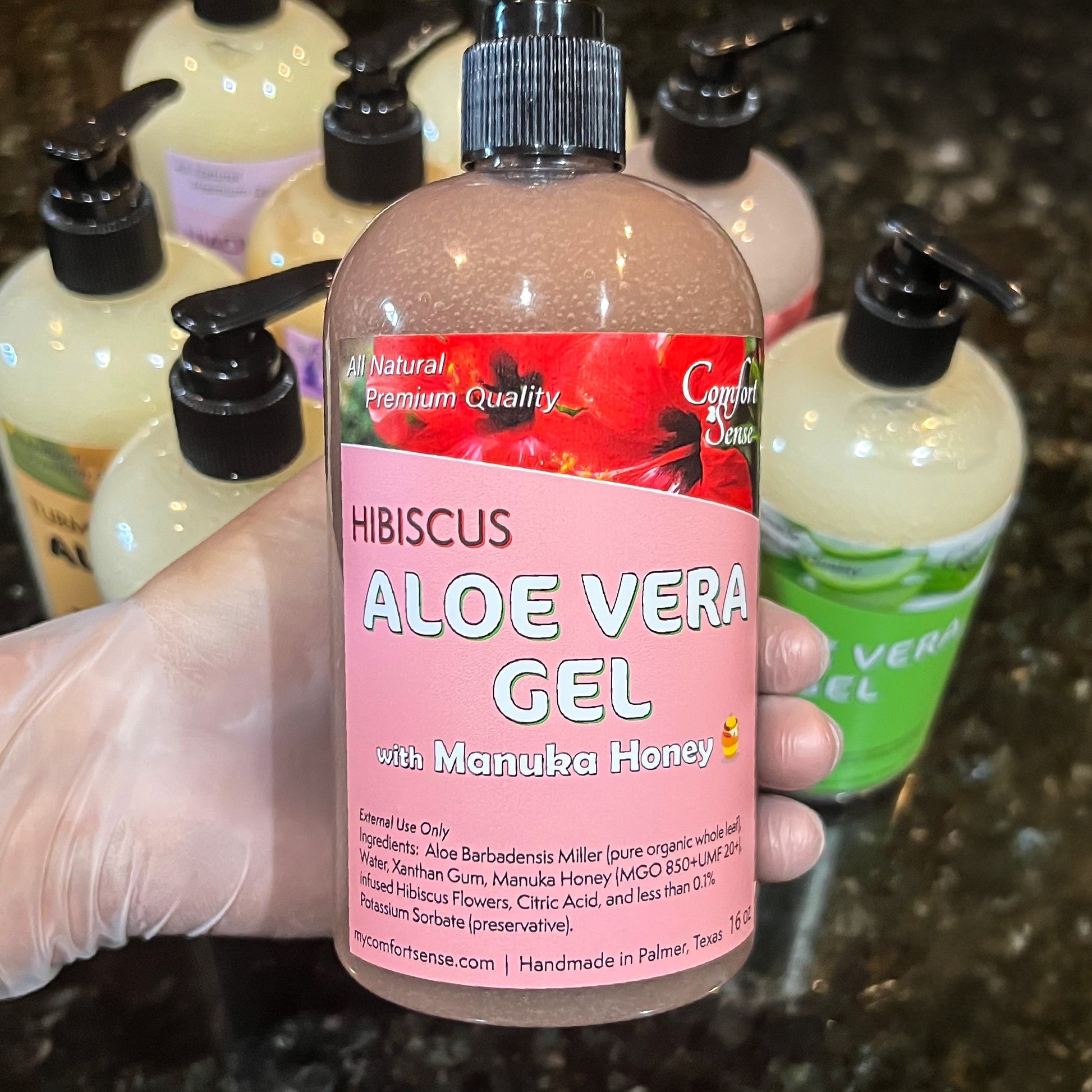 Bottle of hibiscus aloe vera gel with manuka honey held by a gloved hand, surrounded by other similar bottles.